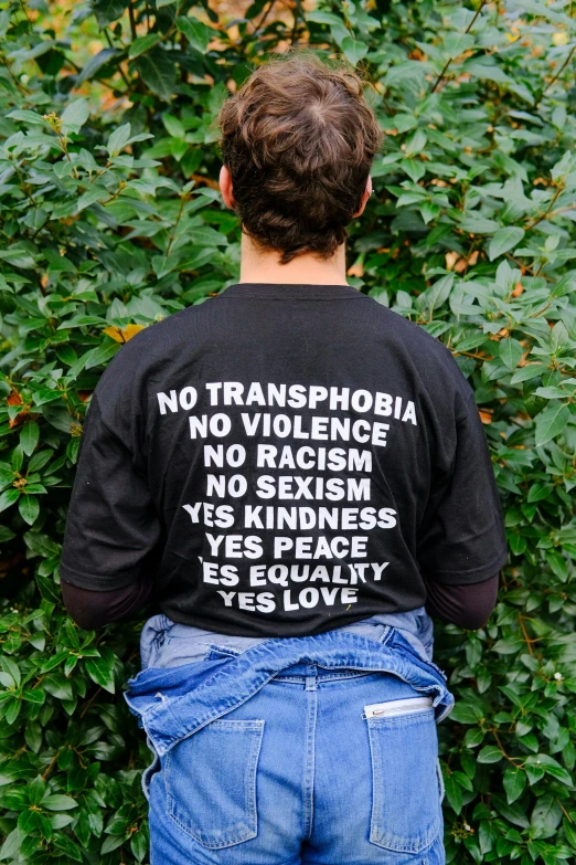 a man wearing a t - shirt that says no transphobia, no violence, no racism, no sexism, yes kindness, yes peace, by Nina Hamnett, photo still of behind view, gardens, black canvas, joe keery