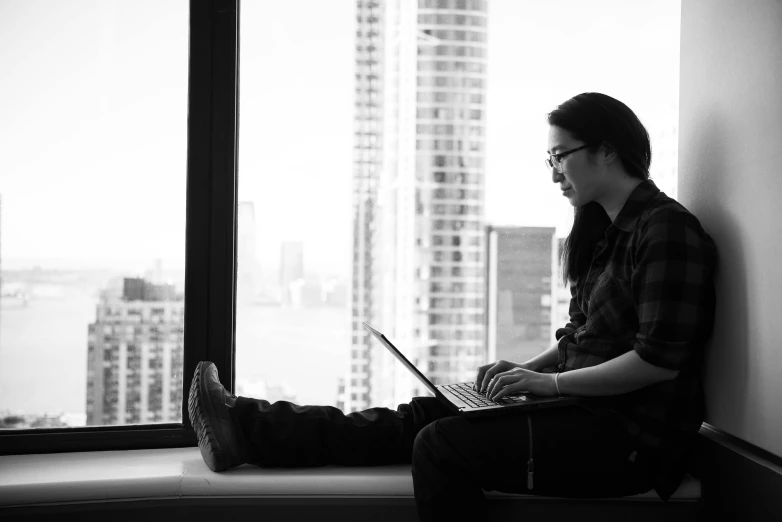 a man sitting on a window sill using a laptop, a black and white photo, inspired by Fei Danxu, developers, david shing, city in background, small chin