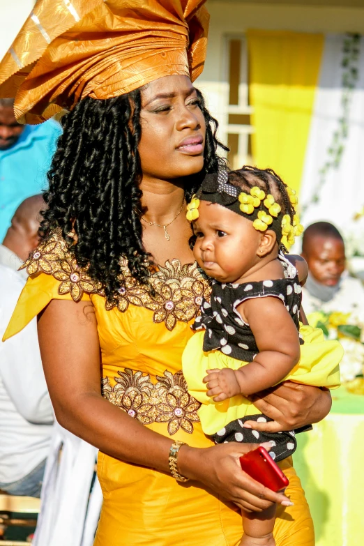 a woman in a yellow dress holding a baby, happening, community celebration, elegantly dressed, dark-skinned, slide show