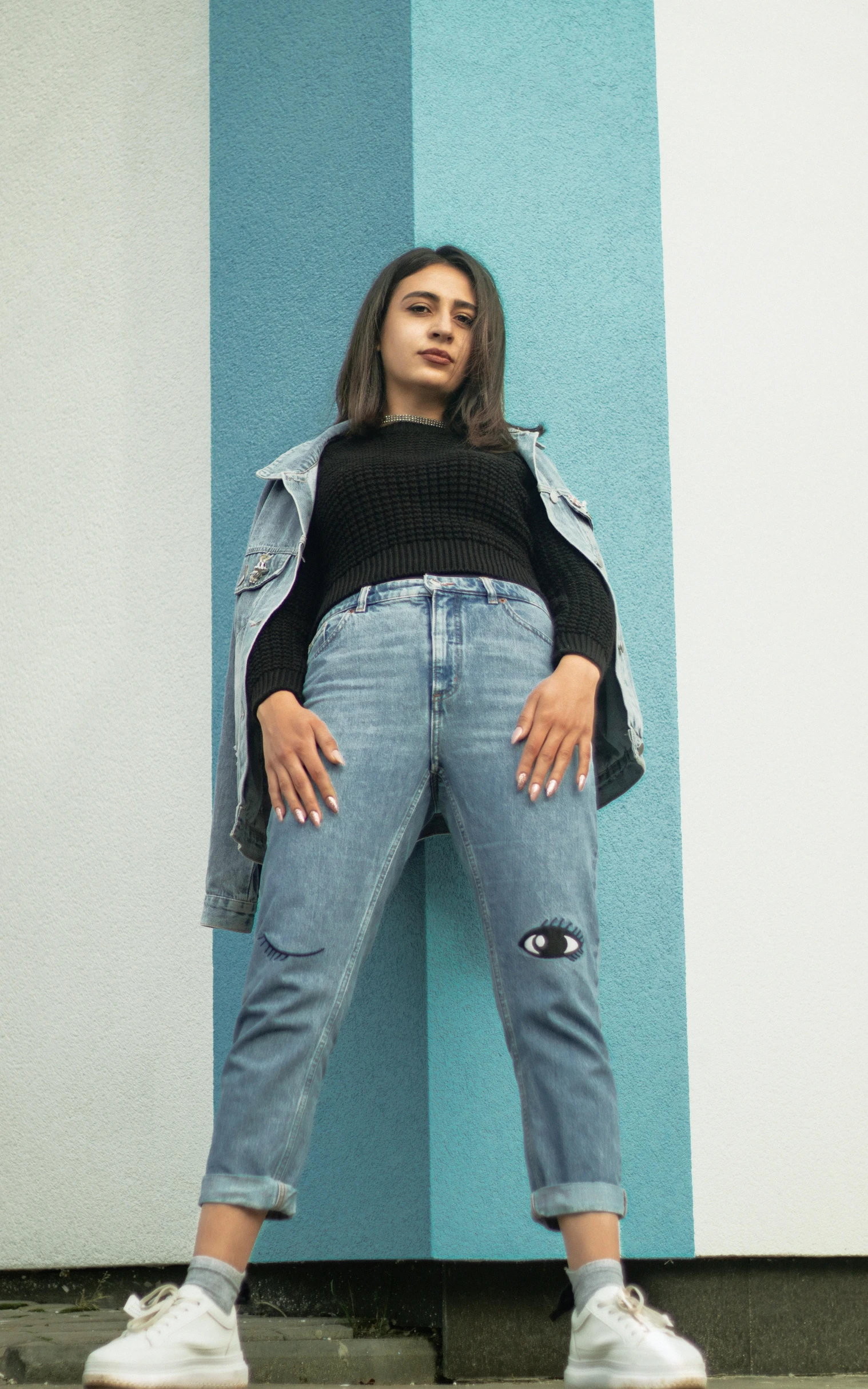 a woman leaning against a blue and white wall, an album cover, inspired by Amelia Peláez, antipodeans, wearing jeans and a black hoodie, wearing a patch over one eye, wearing cargo pants, vista view