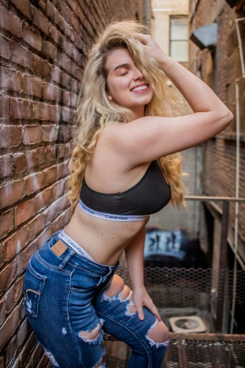 a woman in jeans leaning against a brick wall, a picture, by Niko Henrichon, sport bra, 2 0 yo, bbwchan, curly blond