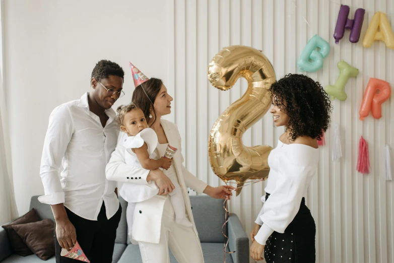 a group of people standing around a birthday cake, white with gold accents, 2 years old, party balloons, thumbnail