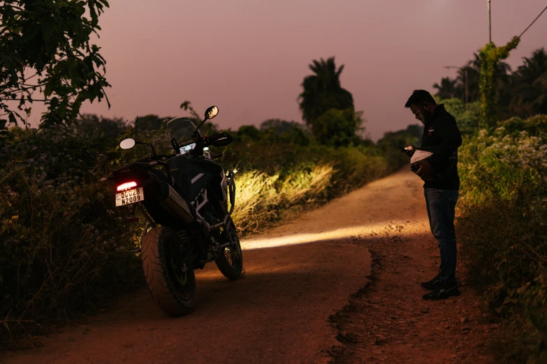 a man standing next to a motorcycle on a dirt road, pexels contest winner, lights on, looking for clues, mechanised, profile image