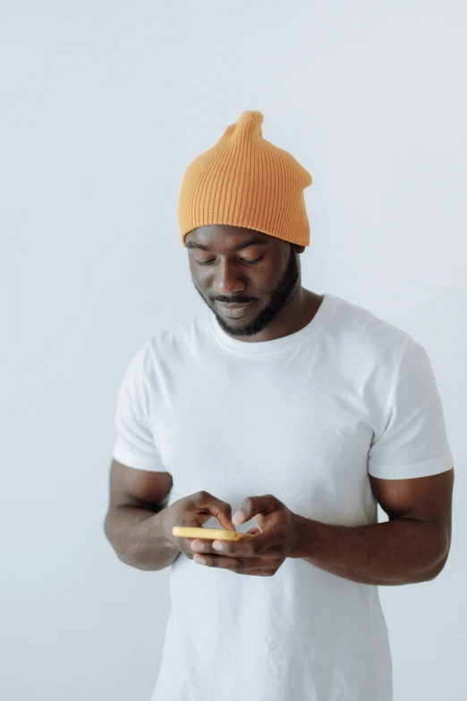 a man in an orange hat is holding a doughnut, trending on pexels, looking at his phone, dark skin tone, beanie hat, dressed in a white t shirt