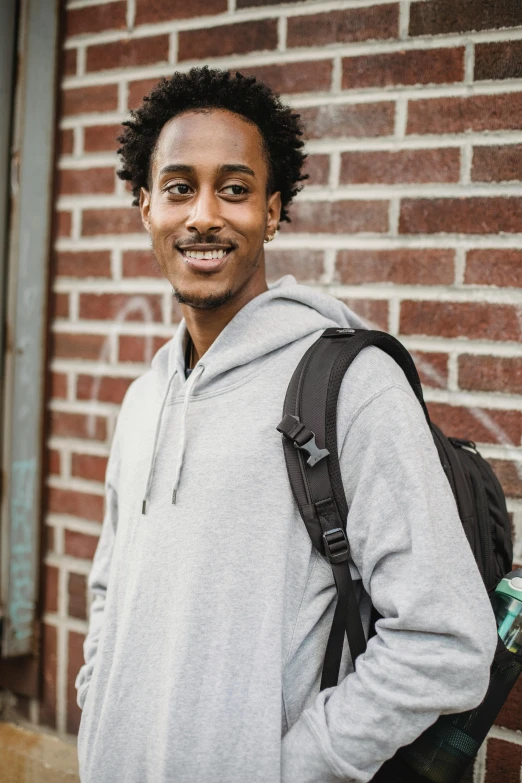 a man standing in front of a brick wall, a man wearing a backpack, gray hoodie, ryan kiera armstrong, loosely cropped