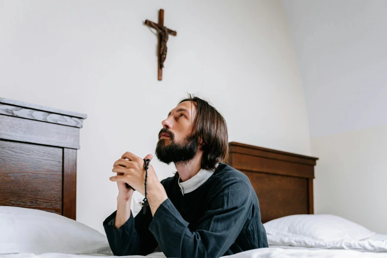 a man sitting on top of a bed next to a cross, a photo, pexels, catholic icon, profile image, looking from shoulder, title