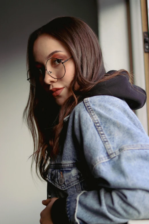 a woman wearing glasses and a denim jacket, a picture, by Robbie Trevino, trending on pexels, charli bowater, brown haired, promotional image, wearing jeans and a black hoodie