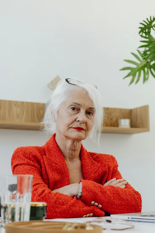 a woman sitting at a table with her arms crossed, by Matija Jama, pexels contest winner, modernism, silver haired, wearing red jacket, sitting on a store shelf, on a white table