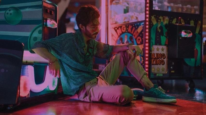 a man sitting on the floor in front of a game machine, inspired by Liam Wong, trending on pexels, magic realism, colourful clothing, hozier, jukebox, iridescent aesthetic