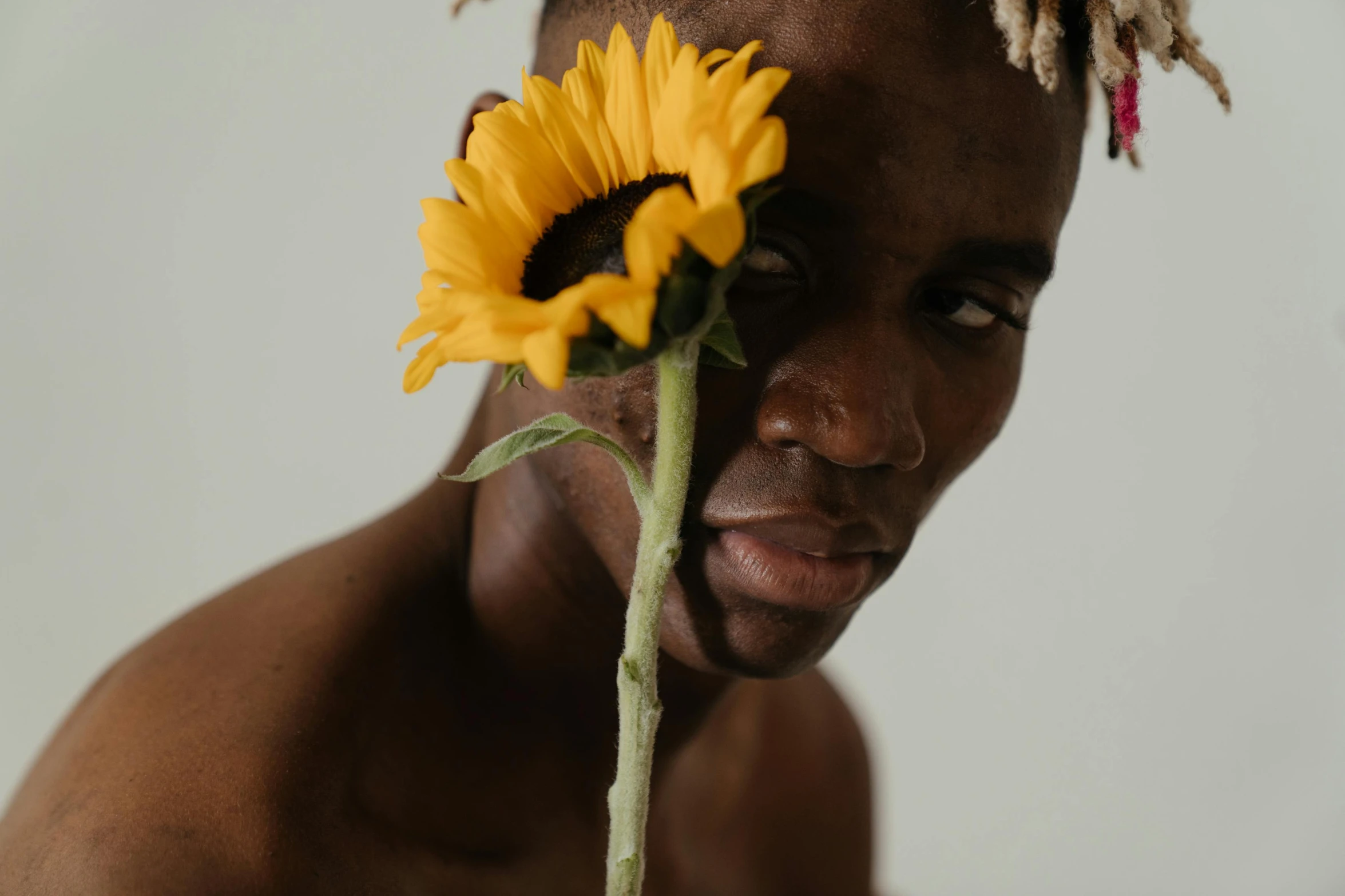 a man with dreadlocks holding a sunflower in front of his face, inspired by Robert Mapplethorpe, trending on pexels, non binary model, with brown skin, tyler the creator, attractive face and body