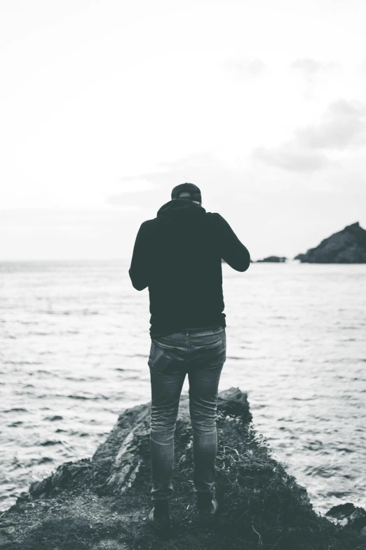 a man standing on top of a rock next to the ocean, a black and white photo, unsplash, happening, wearing jeans and a black hoodie, facing away, profile picture, slightly pixelated