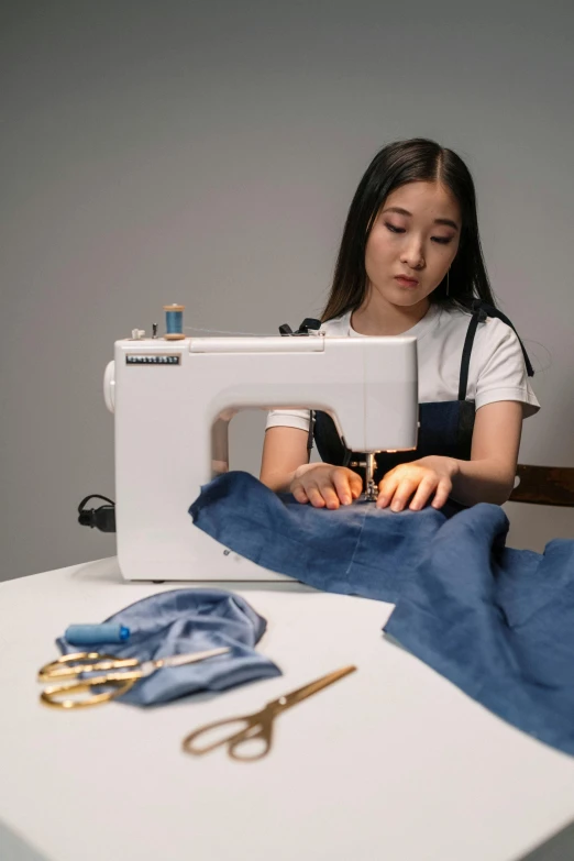 a woman sitting at a table working on a sewing machine, inspired by Ruth Jên, blue clothing, janice sung, made of fabric, product introduction photo