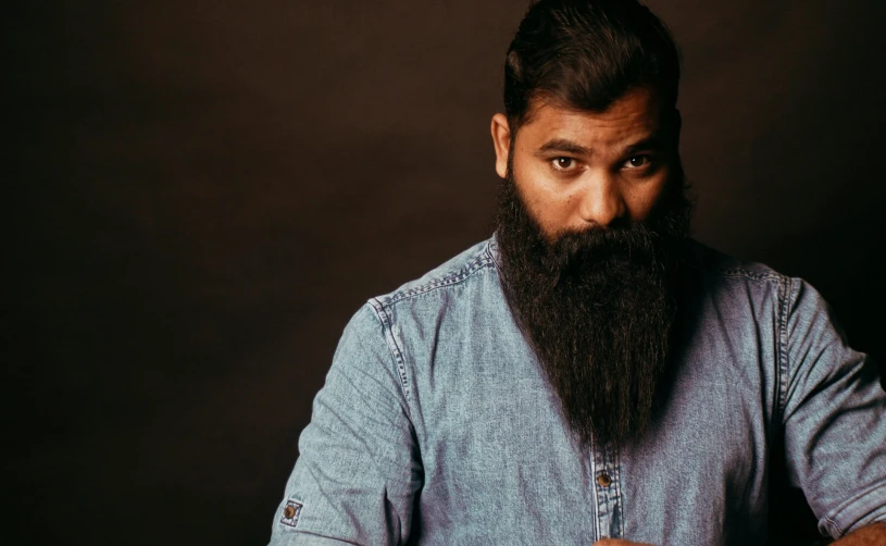 a man with a long beard sitting at a table, pexels contest winner, hurufiyya, wearing a shirt and a jean, solid background, headshot photo, indian