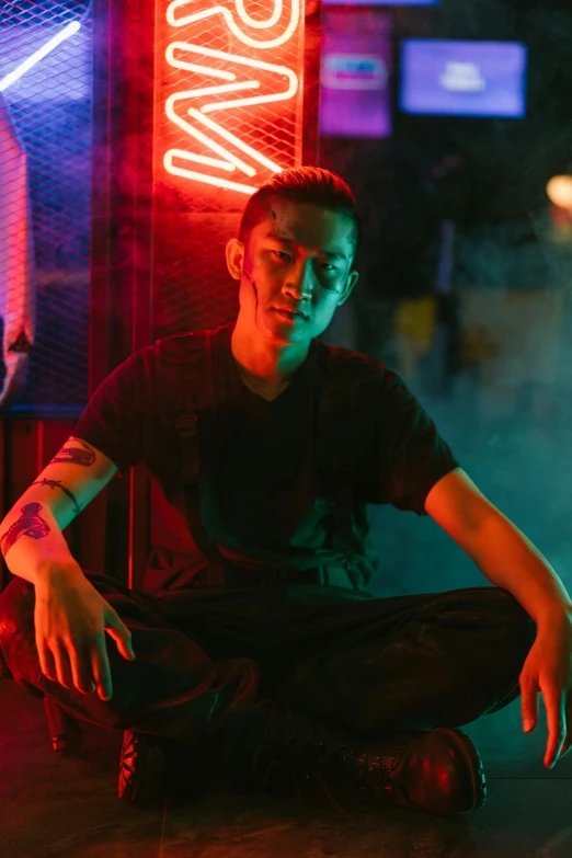 a man sitting on the ground in front of a neon sign, inspired by Liam Wong, trending on pexels, he is wearing a black t-shirt, non binary model, like andy lau, serious lighting