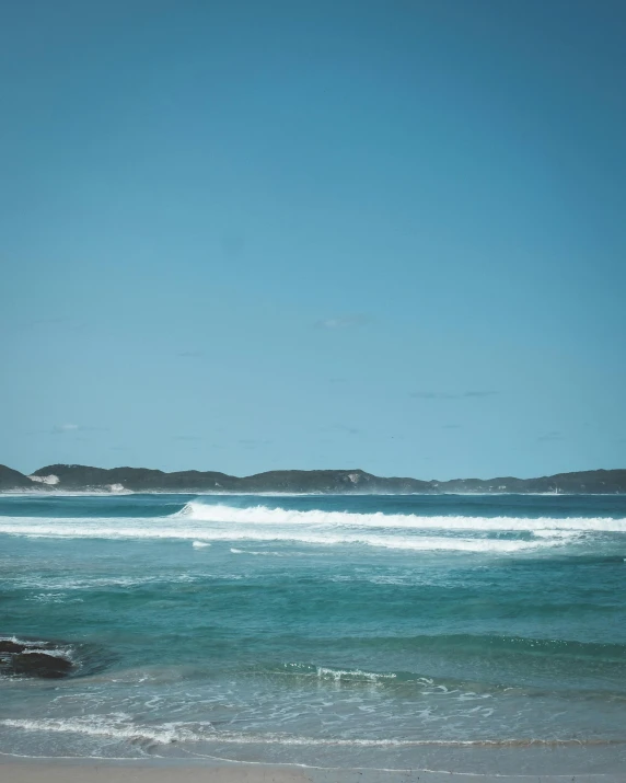 a man riding a surfboard on top of a sandy beach, from the distance, two medium sized islands, blueish, bulli