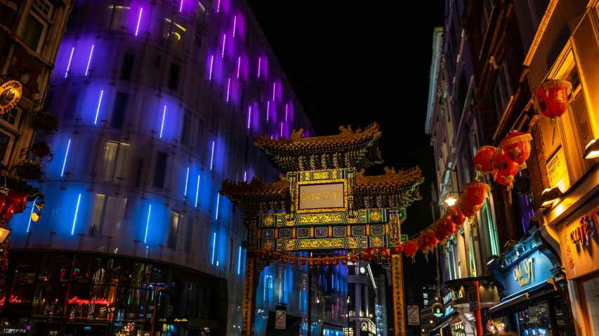 a group of people walking down a street next to tall buildings, unsplash, art nouveau, purple and yellow lighting, chinese building, an archway, london at night