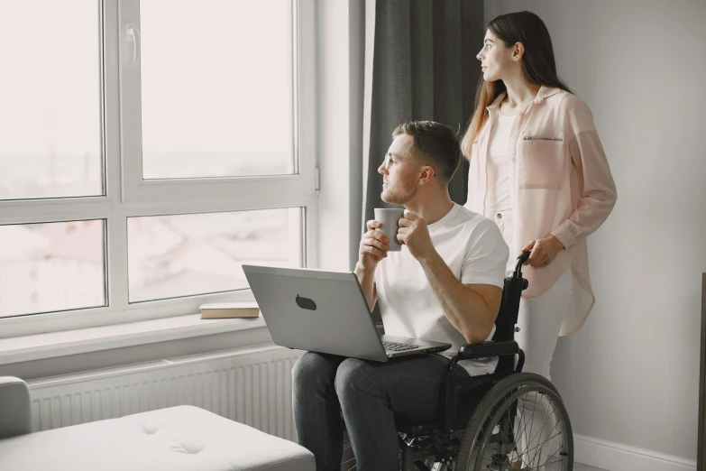 a man in a wheelchair using a laptop next to a woman, by Adam Marczyński, trending on pexels, hurufiyya, standing near a window, lachlan bailey, looking forward, nursing