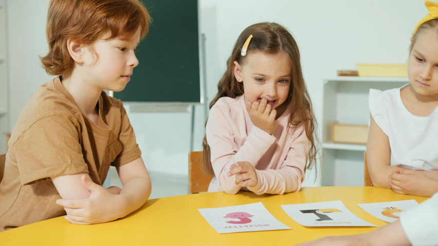 a group of children sitting around a yellow table, trading card game, thumbnail, abcdefghijklmnopqrstuvwxyz, two girls