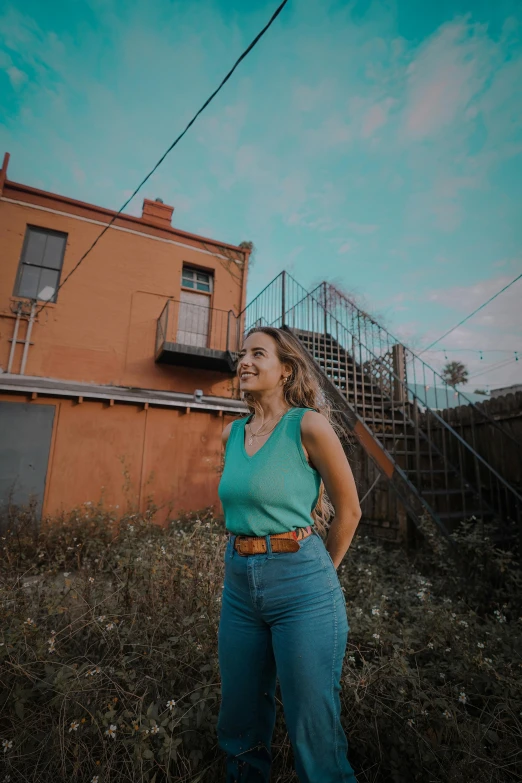 a woman standing in front of a building, teal sky, wearing a shirt and a jean, in savannah, pokimane