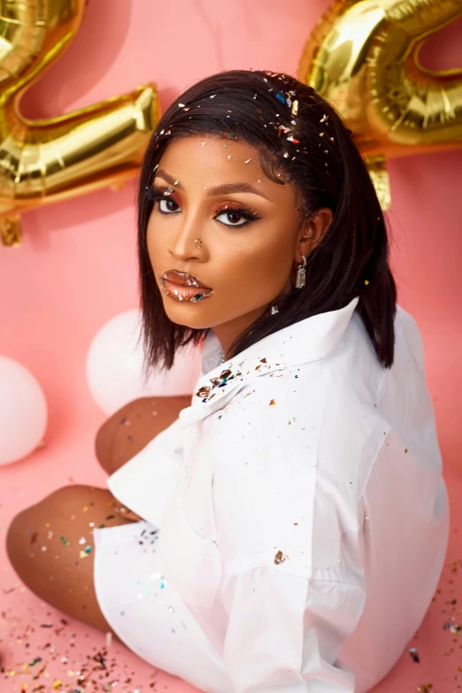 a woman sitting in front of balloons and confetti, an album cover, trending on pexels, visual art, brown skin. light makeup, wearing a white button up shirt, with black glossy lips, official store photo