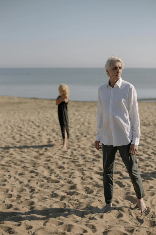 a man standing on top of a sandy beach, an album cover, white haired lady, nonbinary model, older woman, tense