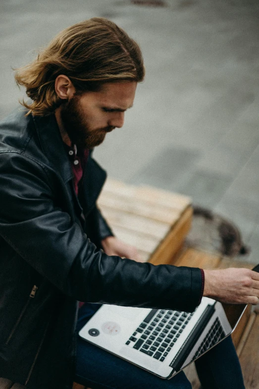 a man sitting on a bench using a laptop, by Jan Tengnagel, trending on unsplash, with a beard and a black jacket, mid long hair, wearing leather, thumbnail