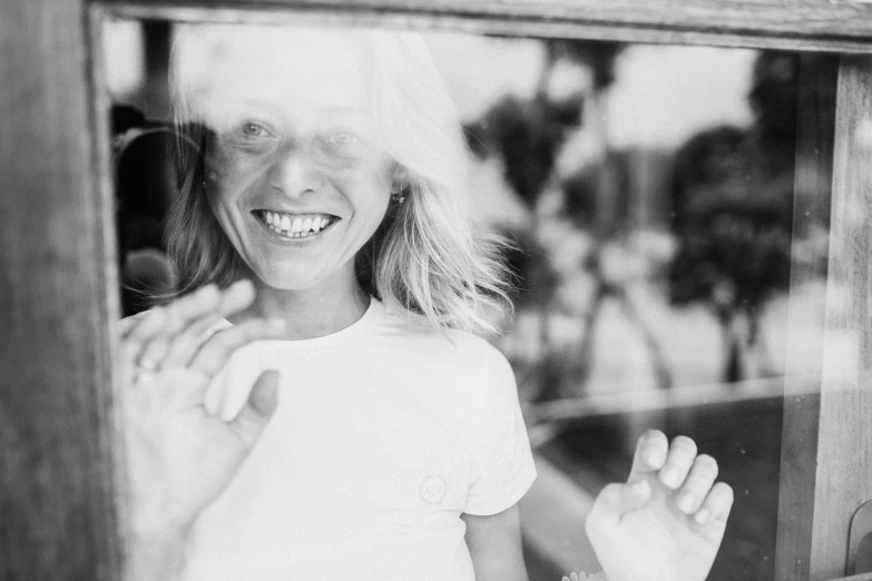 a black and white photo of a little girl looking through a window, by Emma Andijewska, smiling woman, dressed in a white t shirt, a blond, person made out of glass