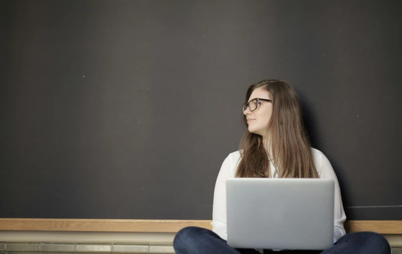 a woman sitting on the floor with a laptop, by Carey Morris, pixabay, blackboard in background, looking across the shoulder, minimal background, background image