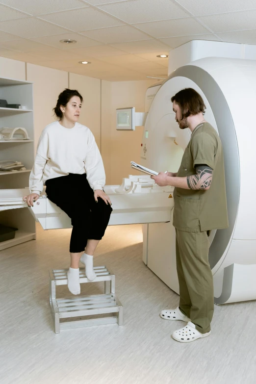 a man standing next to a woman in a room, a hologram, unsplash, medical equipment, sitting on a stool, mri, brown