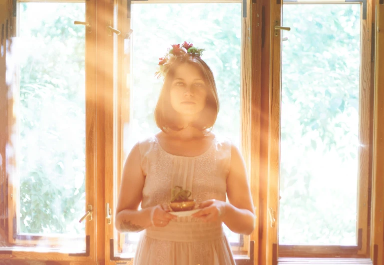 a woman standing in front of a window holding a plate of food, pexels contest winner, light and space, lotus floral crown girl, sunflare, holy ceremony, warmly lit