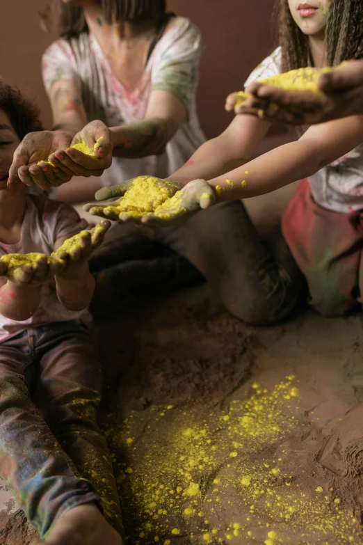 a group of people sitting on top of a pile of dirt, splatter paint, with yellow cloths, kids playing, hands