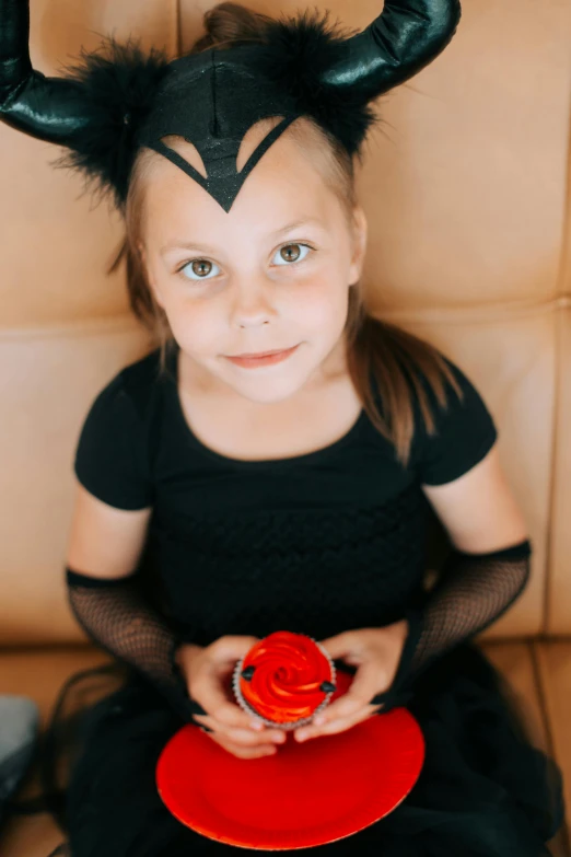 a little girl sitting on a couch holding a red plate, pexels contest winner, wearing black witch hat, frosting on head and shoulders, gif, black cats
