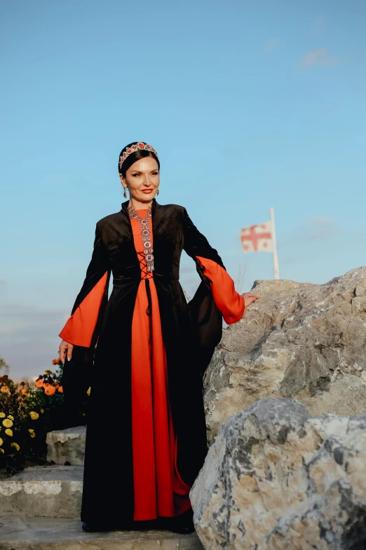 a woman in a black and red dress standing on a rock, wearing long royal robe, georgic, black and orange, wearing authentic attire