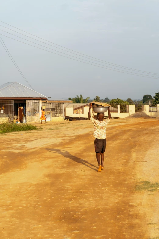 a man walking down a dirt road carrying a basket on his head, by Chinwe Chukwuogo-Roy, happening, fully built buildings, long view, kids playing, exterior