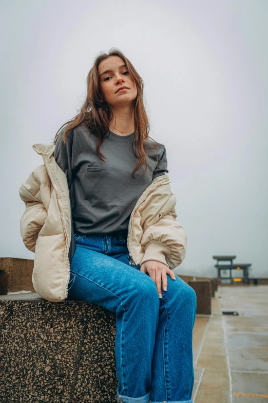 a woman sitting on top of a stone wall, trending on unsplash, happening, wearing sweatshirt, model wears a puffer jacket, jeans and t shirt, natural muted tones