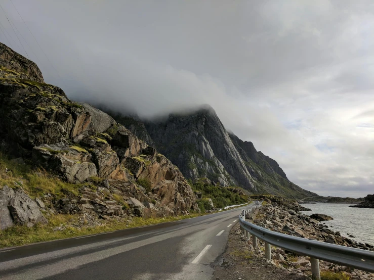 a car driving down a road next to a body of water, by Roar Kjernstad, pexels contest winner, hurufiyya, natural stone road, gray clouds, devils horns, profile image