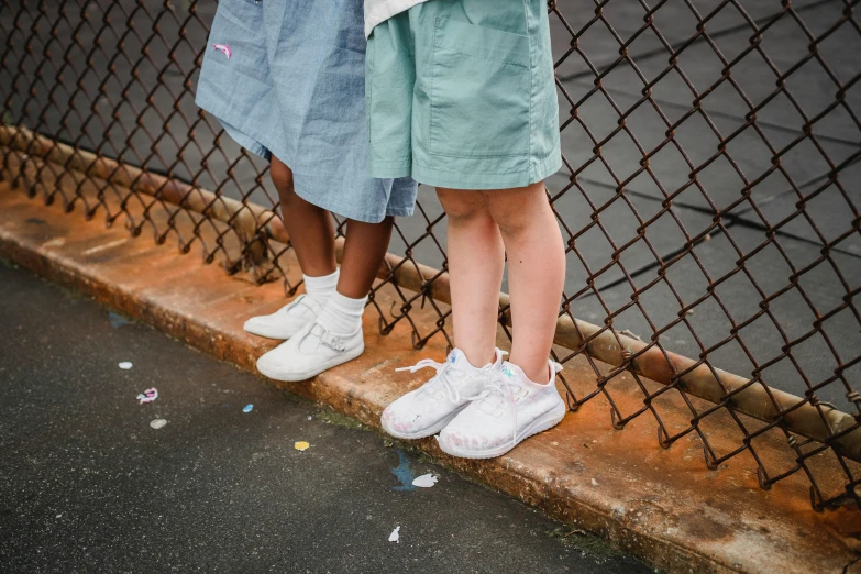 a couple of little girls standing next to a fence, an album cover, by Nina Hamnett, pexels contest winner, wearing white sneakers, detailed shot legs-up, varying ethnicities, triple white colorway