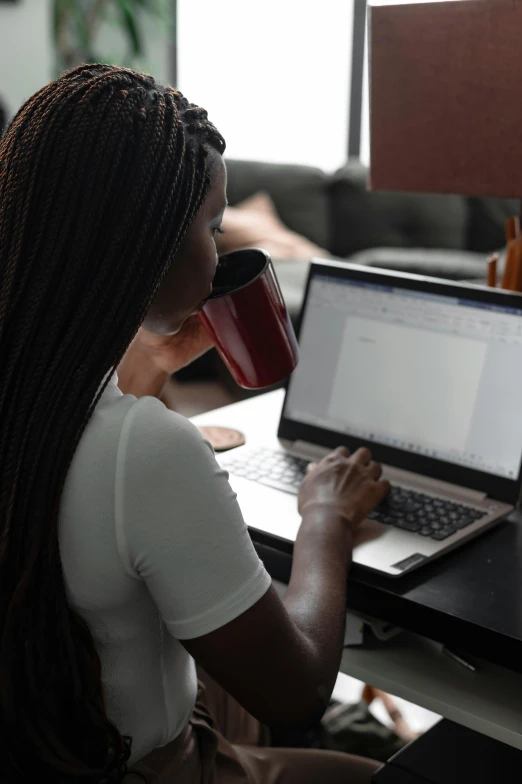 a woman sitting at a desk using a laptop computer, by Carey Morris, pexels contest winner, next to a cup, ( ( dark skin ) ), official screenshot, student