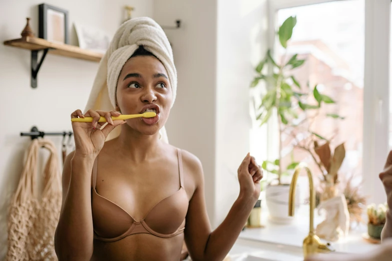 a woman with a towel on her head brushing her teeth, by Emma Andijewska, pexels contest winner, happening, in a yellow bikini, with brown skin, manuka, at home