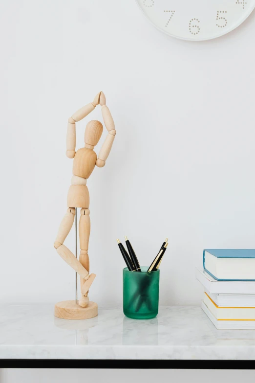 a wooden mannequin sitting on top of a table next to books and a clock, dynamic dancing pose, holding pencil, 2019 trending photo, 12in action figure