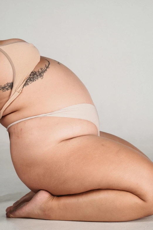 a woman in a bikini sitting on the floor, an album cover, by Matija Jama, trending on pexels, massurrealism, pregnant belly, zoomed in, tattooed body, diaper-shaped