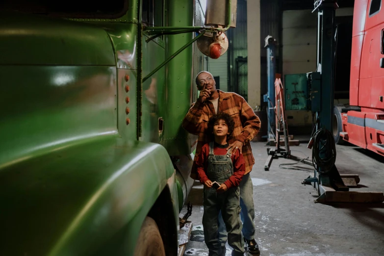 a man standing next to a little boy in front of a green truck, by Dan Frazier, pexels contest winner, in a workshop, francis ford coppola, wearing overalls, charli bowater and artgeem