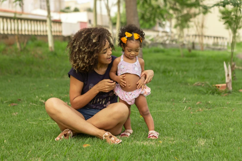 a woman holding a baby sitting on top of a lush green field, a portrait, by Samuel Silva, pexels, figuration libre, african american girl, são paulo, sitting on the ground, high-quality photo
