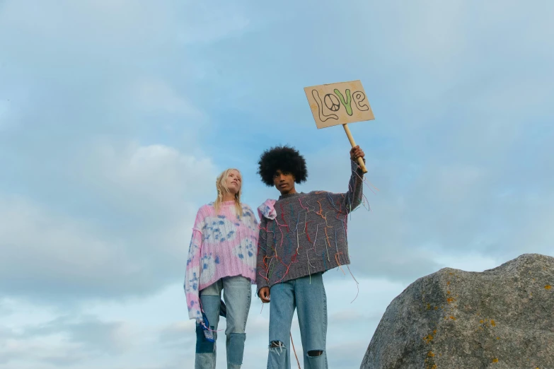 two women standing on top of a rock holding a sign, unsplash, graffiti, flower power, pastel clothing, male and female, product introduction photo