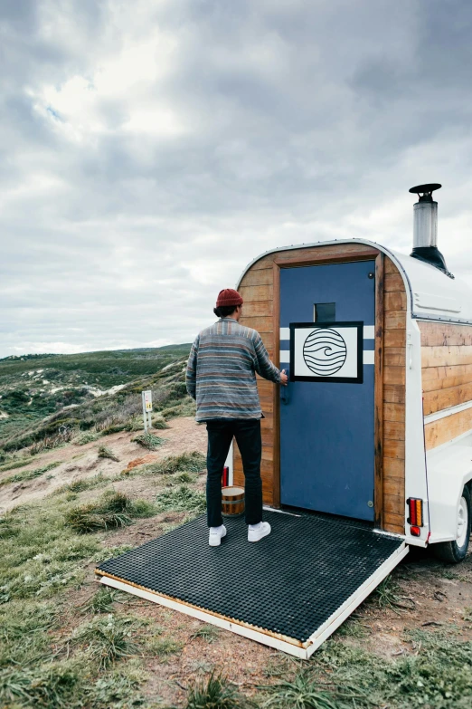 a man standing in front of a trailer with a blue door, trending on unsplash, private press, overlooking the ocean, open vault, built on a steep hill, views front side and rear