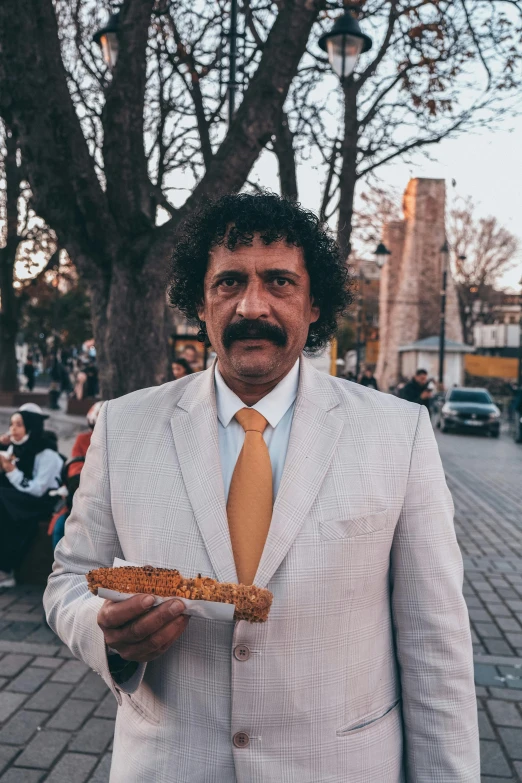 a man in a white suit holding a piece of food, inspired by Osman Hamdi Bey, pexels contest winner, downtown mexico, wearing a worn out brown suit, istanbul, long and orange mustache