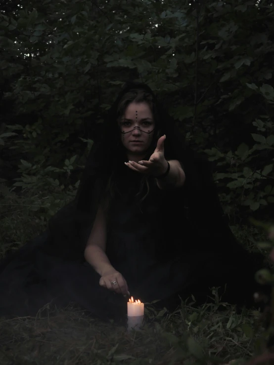 a woman sitting in the grass holding a lit candle, an album cover, by Attila Meszlenyi, wearing black wizard robes, forming a burning hand spell, with haunted eyes and glasses, in a dark forest low light