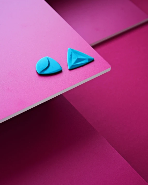 a pair of blue earrings sitting on top of a pink surface, inspired by Frederick Hammersley, trending on pexels, plasticien, normal map, shaped like a yacht, material design, close - up photograph