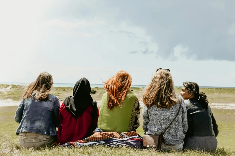 a group of women sitting on top of a grass covered field, a picture, by Alice Mason, trending on pexels, hurufiyya, looking out over the sea, profile image, picnic, mix of ethnicities and genders