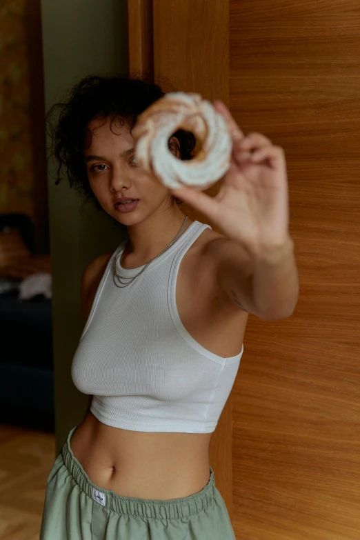 a woman holding a donut in front of her face, an album cover, inspired by Nan Goldin, trending on pexels, renaissance, white tank top, barbara palvin, physical : tinyest midriff ever, tessa thompson inspired